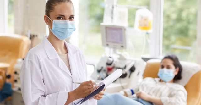 Portrait of young female doctor in mask writing in medical card and looking at camera with patient in the background